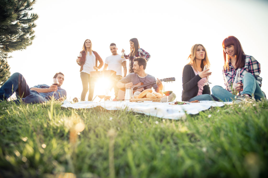 Group of friends picnicking in the grass at a park, laughing while one plays the guitar